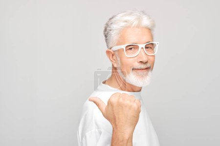 Foto de Hombre maduro de pelo gris 60 años de edad en camiseta blanca señalando a un lado el dedo, demostrando el espacio vacío para el producto o el texto aislado en el fondo del estudio blanco - Imagen libre de derechos