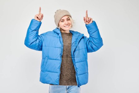 Téléchargez les photos : Jeune femme veste d'hiver bleue pointant du doigt, démontrant l'espace vide pour le produit ou le texte isolé sur backgroun studio blanc - en image libre de droit