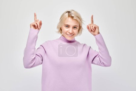 Téléchargez les photos : Jeune femme blonde au doigt pointant occasionnellement vers le haut, démontrant un espace vide pour le produit ou le texte isolé sur fond de studio blanc - en image libre de droit