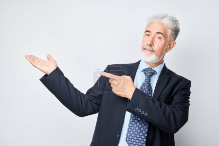 Foto de Hombre maduro de pelo gris de 60 años con traje de negocios sosteniendo algo en la mano, demostrando espacio vacío para el producto o el texto aislado en el fondo del estudio blanco - Imagen libre de derechos