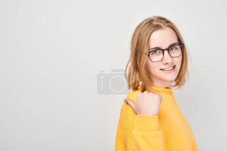 Téléchargez les photos : Jeune fille blonde en jaune casual pointant le doigt de côté, démontrant un espace vide pour le produit ou le texte isolé sur fond de studio blanc - en image libre de droit