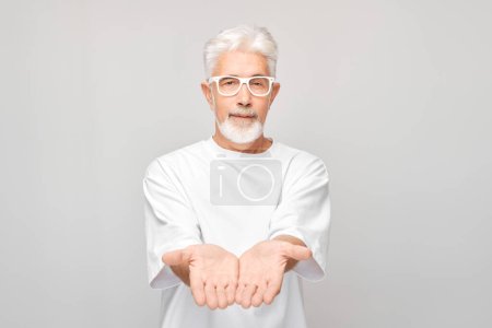 Foto de Hombre maduro de pelo gris de 60 años con camiseta blanca sosteniendo algo en la mano, demostrando espacio vacío para el producto o el texto aislado en el fondo del estudio blanco - Imagen libre de derechos