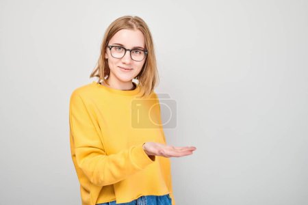 Foto de Jovencita rubia en amarillo casual sosteniendo algo en la mano, demostrando espacio vacío para producto o texto aislado en fondo de estudio blanco - Imagen libre de derechos