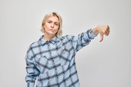 Photo for Portrait of blond girl in plaid shirt showing thumbs down isolated on white studio background. Dislike, wrong choice, negative evaluation concep - Royalty Free Image