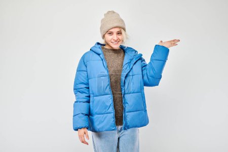 Foto de Mujer joven con chaqueta azul de invierno sosteniendo algo en la mano, demostrando espacio vacío para el producto o el texto aislado en el fondo del estudio blanco - Imagen libre de derechos
