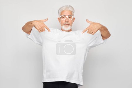 Photo for Gray-haired mature man 60 years old in white T-shirt pointing finger down, demonstrating empty space for product or text isolated on white studio backgroun - Royalty Free Image