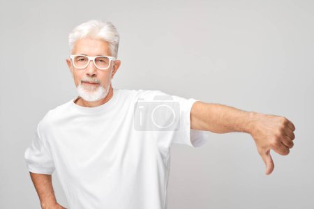 Téléchargez les photos : Portrait d'un homme âgé aux cheveux gris montrant les pouces vers le bas, isolé sur fond de studio blanc. Refus, mauvais choix, concept d'évaluation négative - en image libre de droit