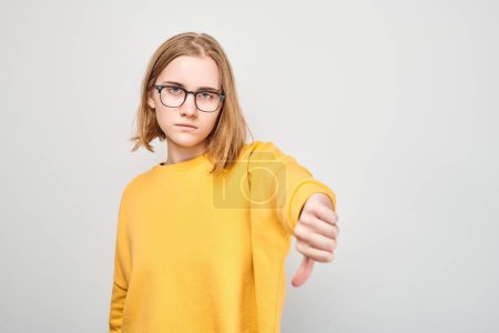 Téléchargez les photos : Portrait de jeune étudiante en lunettes et cardigan jaune montrant les pouces vers le bas isolés sur fond de studio blanc. Refus, mauvais choix, évaluation négative concéder - en image libre de droit