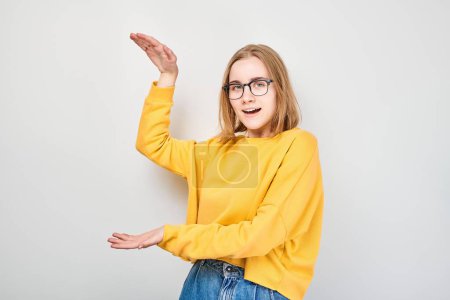 Téléchargez les photos : Jeune fille blonde en jaune casual tenant quelque chose à la main, démontrant un espace vide pour le produit ou le texte isolé sur fond de studio blanc - en image libre de droit