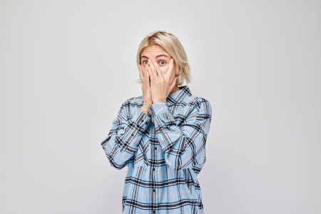 Téléchargez les photos : Portrait de jeune femme blonde couvrant les yeux avec la main, regardant à travers les doigts isolés sur fond de studio blanc - en image libre de droit
