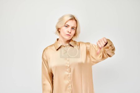 Photo for Portrait of blond girl in casual shirt showing thumbs down isolated on white studio background. Dislike, wrong choice, negative evaluation concep - Royalty Free Image