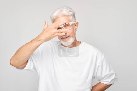 Téléchargez les photos : Portrait d'homme âgé couvrant les yeux avec la main, regardant à travers les doigts isolés sur fond blanc studio - en image libre de droit