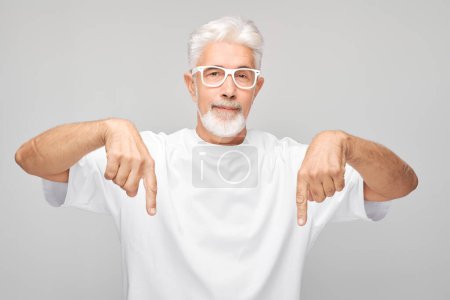 Photo for Gray-haired mature man 60 years old in white T-shirt pointing finger down, demonstrating empty space for product or text isolated on white studio backgroun - Royalty Free Image