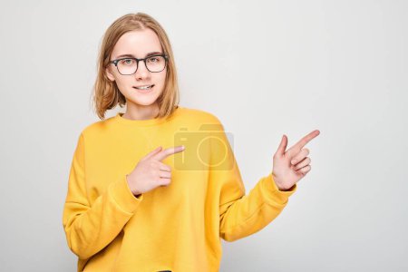 Téléchargez les photos : Jeune fille blonde en jaune casual pointant le doigt de côté, démontrant un espace vide pour le produit ou le texte isolé sur fond de studio blanc - en image libre de droit