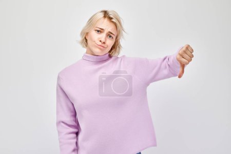 Téléchargez les photos : Portrait de fille blonde en pull lilas montrant pouces vers le bas isolé sur fond de studio blanc. Refus, mauvais choix, évaluation négative concéder - en image libre de droit
