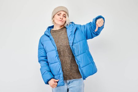 Photo for Portrait of blond girl in blue winter clothes showing thumbs down isolated on white studio background. Dislike, wrong choice, negative evaluation concept - Royalty Free Image