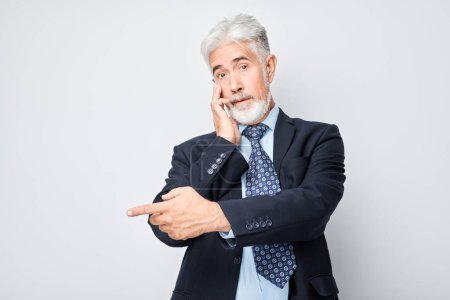 Foto de Hombre maduro de pelo gris 60 años en traje de negocios señalando a un lado el dedo, demostrando espacio vacío para el producto o el texto aislado en el fondo del estudio blanco - Imagen libre de derechos