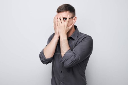 Photo for Portrait of young businessman covering eyes with hand, peeking through fingers isolated on white studio backgroun - Royalty Free Image