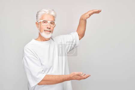 Téléchargez les photos : Homme mûr aux cheveux gris 60 ans en T-shirt blanc tenant quelque chose à la main, démontrant un espace vide pour le produit ou le texte isolé sur un backgroun studio blanc - en image libre de droit