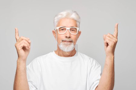 Photo for Gray-haired mature man 60 years old in white T-shirt pointing up finger, demonstrating empty space for product or text isolated on white studio backgroun - Royalty Free Image