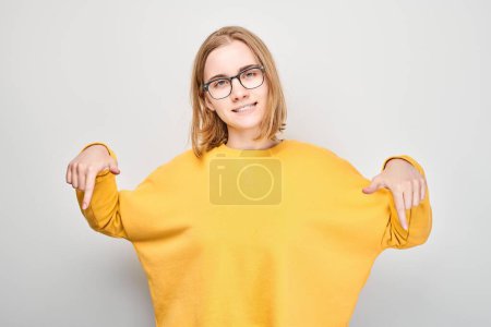 Téléchargez les photos : Jeune fille blonde en jaune casual pointant le doigt vers le bas, démontrant un espace vide pour le produit ou le texte isolé sur fond de studio blanc - en image libre de droit