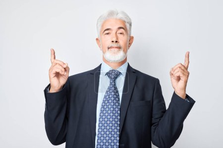 Foto de Hombre maduro de pelo gris de 60 años con traje de negocios apuntando hacia arriba con el dedo, demostrando espacio vacío para el producto o el texto aislado en el fondo del estudio blanco - Imagen libre de derechos