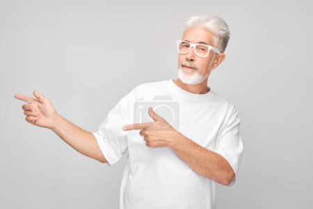 Téléchargez les photos : Homme mûr aux cheveux gris 60 ans en T-shirt blanc pointant du doigt, démontrant un espace vide pour le produit ou le texte isolé sur un backgroun studio blanc - en image libre de droit