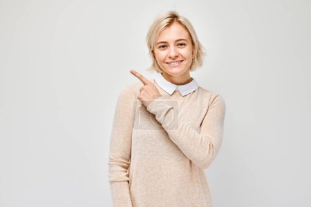 Téléchargez les photos : Jeune femme blonde au doigt pointant vers l'extérieur, démontrant un espace vide pour le produit ou le texte isolé sur fond de studio blanc - en image libre de droit