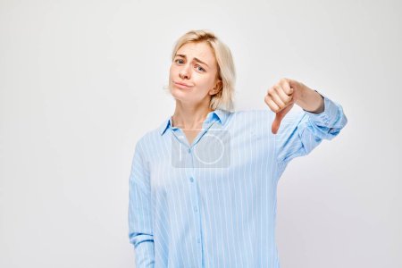 Foto de Retrato de una chica rubia con camisa casual que muestra los pulgares hacia abajo aislados sobre fondo blanco del estudio. Odio, elección equivocada, evaluación negativa concep - Imagen libre de derechos