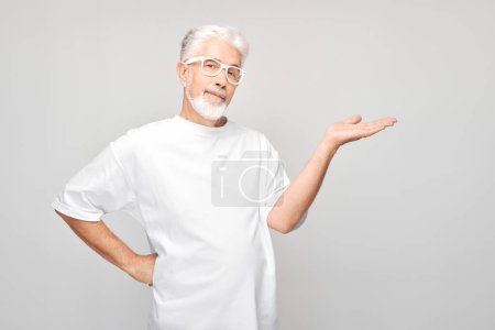 Photo for Gray-haired mature man 60 years old in white T-shirt holding something in hand, demonstrating empty space for product or text isolated on white studio backgroun - Royalty Free Image