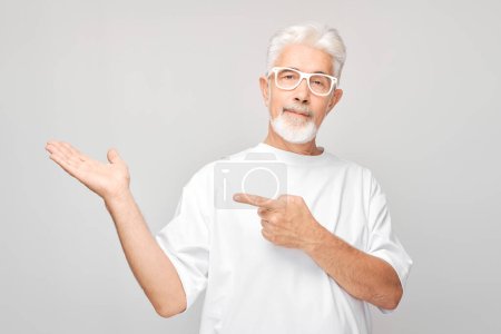 Photo for Gray-haired mature man 60 years old in white T-shirt holding something in hand, demonstrating empty space for product or text isolated on white studio backgroun - Royalty Free Image