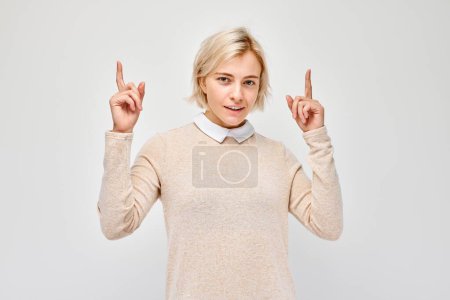 Foto de Mujer rubia joven en casual apuntando con el dedo hacia arriba, demostrando espacio vacío para el producto o el texto aislado en el fondo del estudio blanco - Imagen libre de derechos