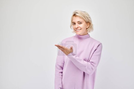 Téléchargez les photos : Jeune femme blonde décontractée tenant quelque chose en main, démontrant un espace vide pour le produit ou le texte isolé sur fond de studio blanc - en image libre de droit