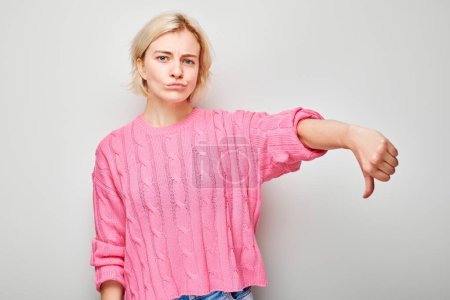 Foto de Retrato de chica rubia en rosa casual mostrando los pulgares hacia abajo aislado sobre fondo blanco del estudio. Odio, elección equivocada, evaluación negativa concep - Imagen libre de derechos