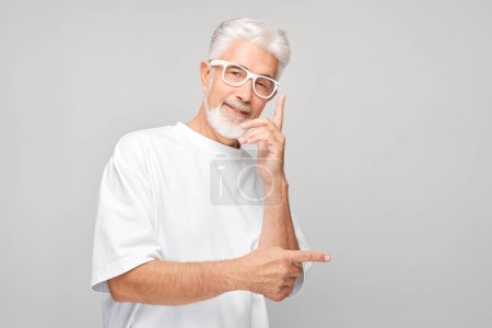 Téléchargez les photos : Homme mûr aux cheveux gris 60 ans en T-shirt blanc pointant du doigt, démontrant un espace vide pour le produit ou le texte isolé sur un backgroun studio blanc - en image libre de droit