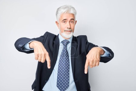 Foto de Hombre maduro de cabello gris de 60 años con traje de negocios apuntando hacia abajo con el dedo, demostrando espacio vacío para el producto o el texto aislado en el fondo del estudio blanco - Imagen libre de derechos