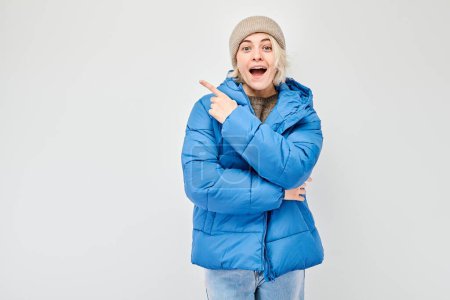 Téléchargez les photos : Jeune femme veste d'hiver bleue pointant du doigt, démontrant l'espace vide pour le produit ou le texte isolé sur fond de studio blanc - en image libre de droit