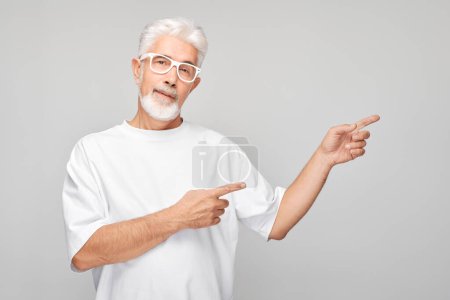 Foto de Hombre maduro de pelo gris 60 años de edad en camiseta blanca señalando a un lado el dedo, demostrando el espacio vacío para el producto o el texto aislado en el fondo del estudio blanco - Imagen libre de derechos