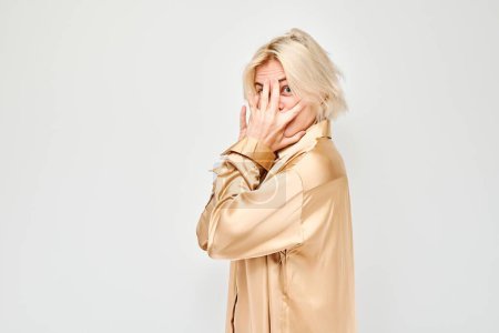 Photo for Portrait of young blonde woman covering eyes with hand, peeking through fingers isolated on white studio backgroun - Royalty Free Image