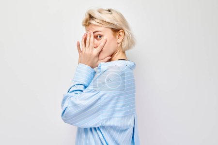 Téléchargez les photos : Portrait de jeune femme blonde couvrant les yeux avec la main, regardant à travers les doigts isolés sur fond de studio blanc - en image libre de droit
