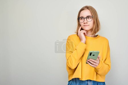Photo for Portrait of young blond woman looks on mobile phone and thinks. Person with smartphone isolated on white backgroun - Royalty Free Image