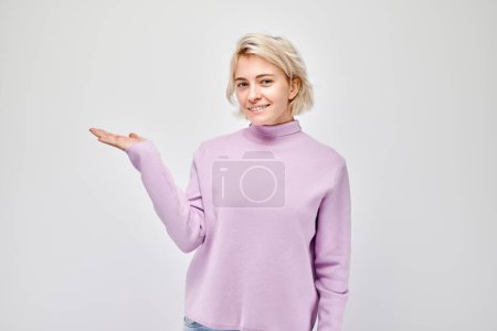 Téléchargez les photos : Jeune femme blonde décontractée tenant quelque chose en main, démontrant un espace vide pour le produit ou le texte isolé sur fond de studio blanc - en image libre de droit