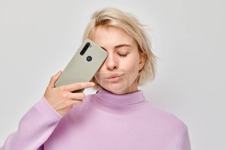 Téléchargez les photos : Portrait de jeune femme blonde prend selfie photo sur téléphone portable. Blogueur avec smartphone isolé sur fond blanc - en image libre de droit