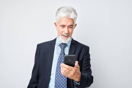 Foto de Retrato de un anciano hombre de negocios con traje que sostiene el teléfono móvil en la mano con la cara sonriente feliz. Persona con teléfono inteligente aislado en el fondo blanco - Imagen libre de derechos