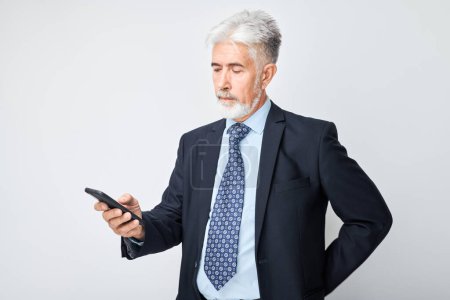 Téléchargez les photos : Portrait d'un homme d'affaires âgé en costume regarde sur le téléphone mobile et pense. Personne avec smartphone isolé sur fond blanc - en image libre de droit