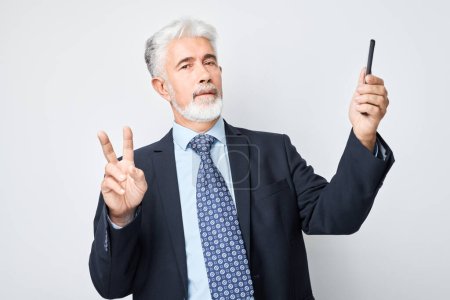 Téléchargez les photos : Portrait d'un homme d'affaires âgé en costume prend selfie photo sur téléphone mobile. Blogueur avec smartphone isolé sur fond blanc - en image libre de droit
