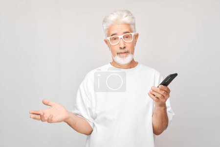 Téléchargez les photos : Portrait d'homme aux cheveux gris en T-shirt blanc regarde sur téléphone portable et pense. Personne avec smartphone isolé sur fond blanc - en image libre de droit
