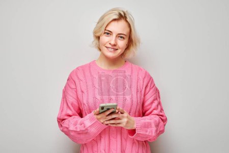 Téléchargez les photos : Portrait de jeune femme blonde en pull rose tenant le téléphone portable à la main avec un visage souriant heureux. Personne avec smartphone isolé sur fond blanc - en image libre de droit