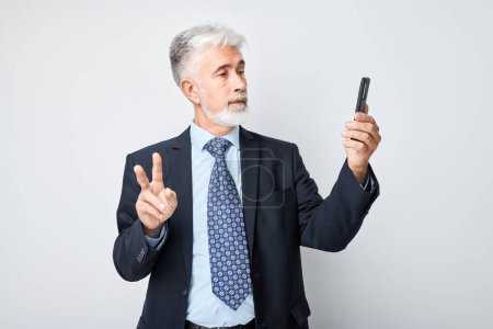 Téléchargez les photos : Portrait d'un homme d'affaires âgé en costume prend selfie photo sur téléphone mobile. Blogueur avec smartphone isolé sur fond blanc - en image libre de droit