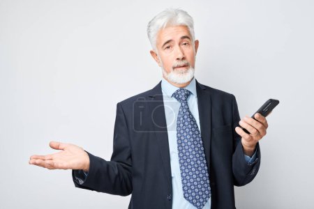Foto de Retrato de hombre de negocios de edad avanzada en traje se ve en el teléfono móvil y piensa. Persona con teléfono inteligente aislado en el fondo blanco - Imagen libre de derechos